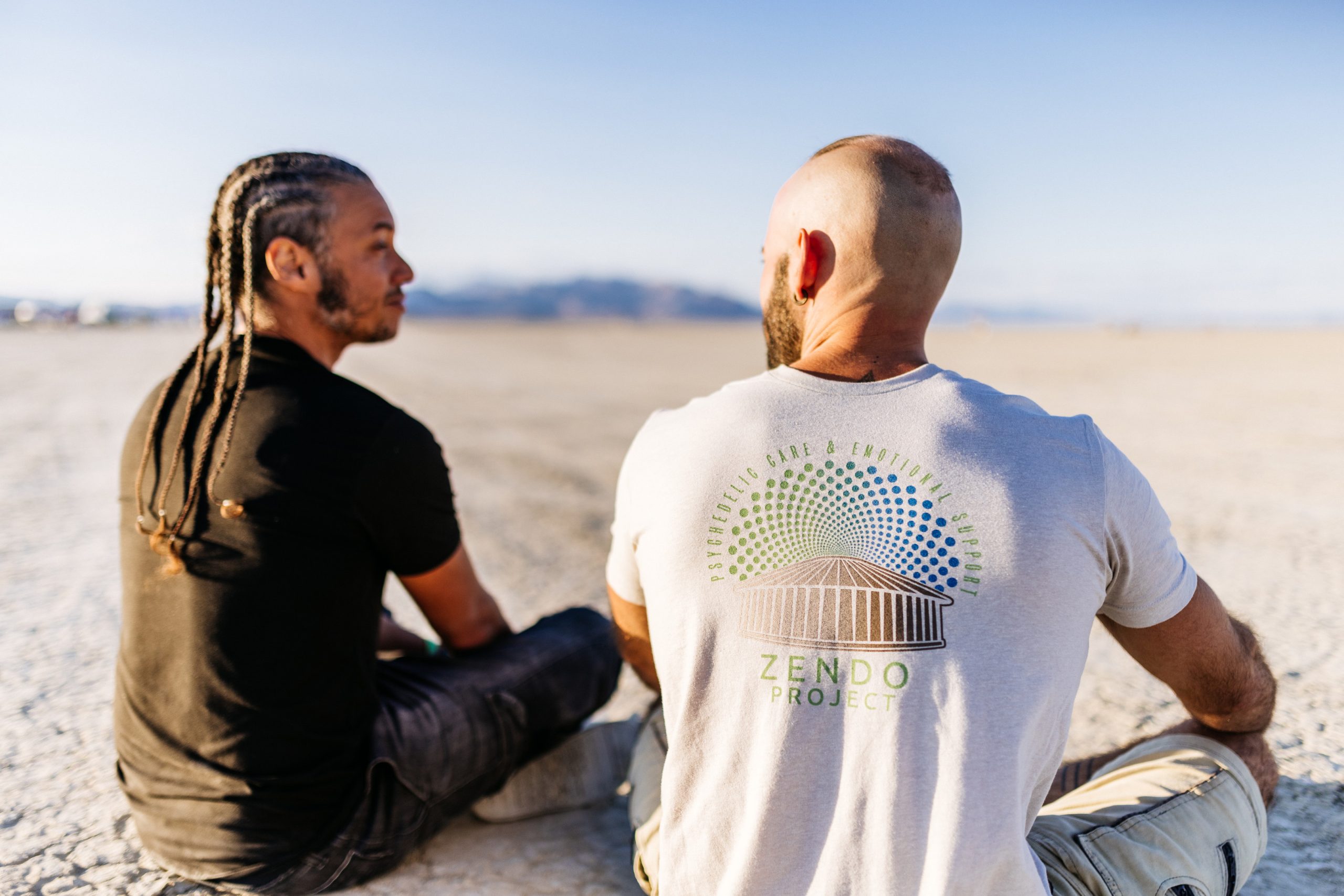 Two people sit next to each other on the playa at Burning Man. The person on the right is wearing a Zendo Project T-shirt.